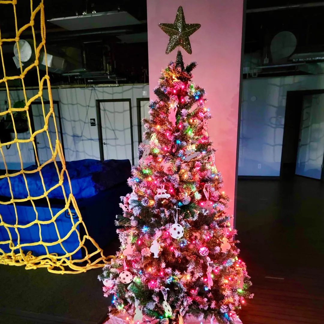 Colorful Christmas tree adorned with lights and ornaments, surrounded by a playful environment with a yellow net structure, emphasizing festive holiday spirit for children with sensory processing challenges.