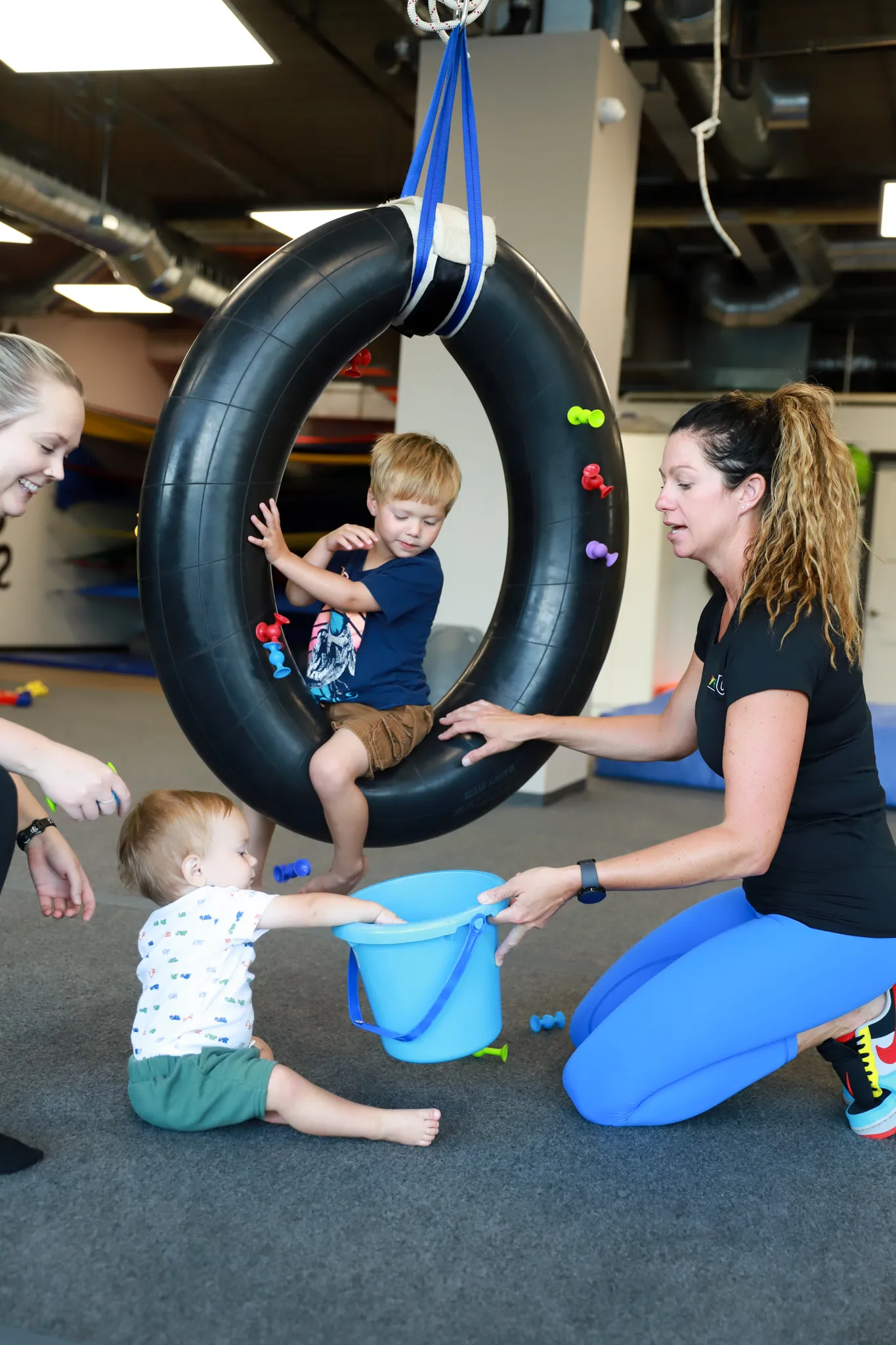 Children engaging in playful activities with therapists, including a boy on a hanging tire swing and a toddler reaching into a blue bucket, promoting developmental skills through play at Move U therapy sessions.