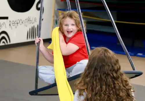 Child on swing engaging in playful therapy session, wearing red shirt and gray pants, with yellow fabric, promoting family involvement in pediatric occupational therapy.