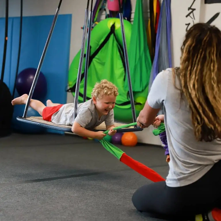 Child joyfully swinging on a therapy swing with clinician support in a pediatric therapy setting, surrounded by sensory equipment, emphasizing child-led play and emotional growth.