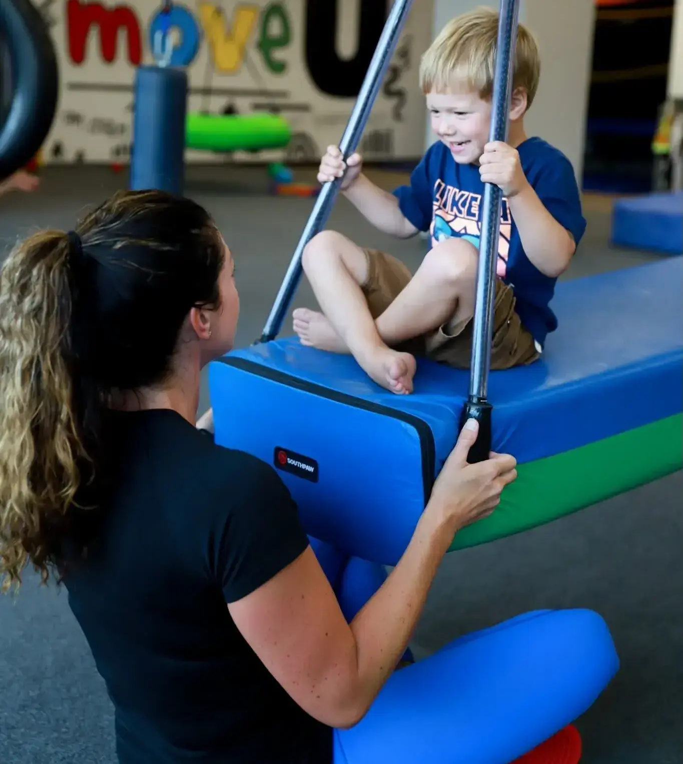 Child enjoying swinging on a padded apparatus, supported by a therapist, in a vibrant pediatric therapy setting promoting emotional growth and sensory integration.