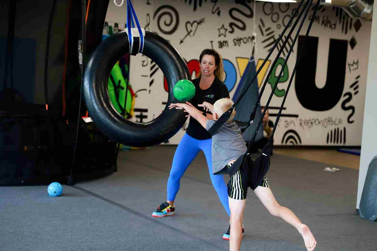 Child engaging in pediatric therapy, reaching for a green ball while suspended in a harness, with a therapist assisting in a colorful therapy space at Move U.